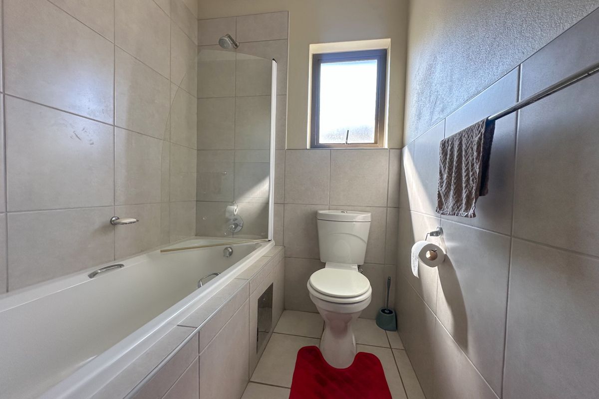 A clean and functional bathroom featuring a built-in bathtub with a glass shower screen, a toilet, and neutral wall and floor tiling.