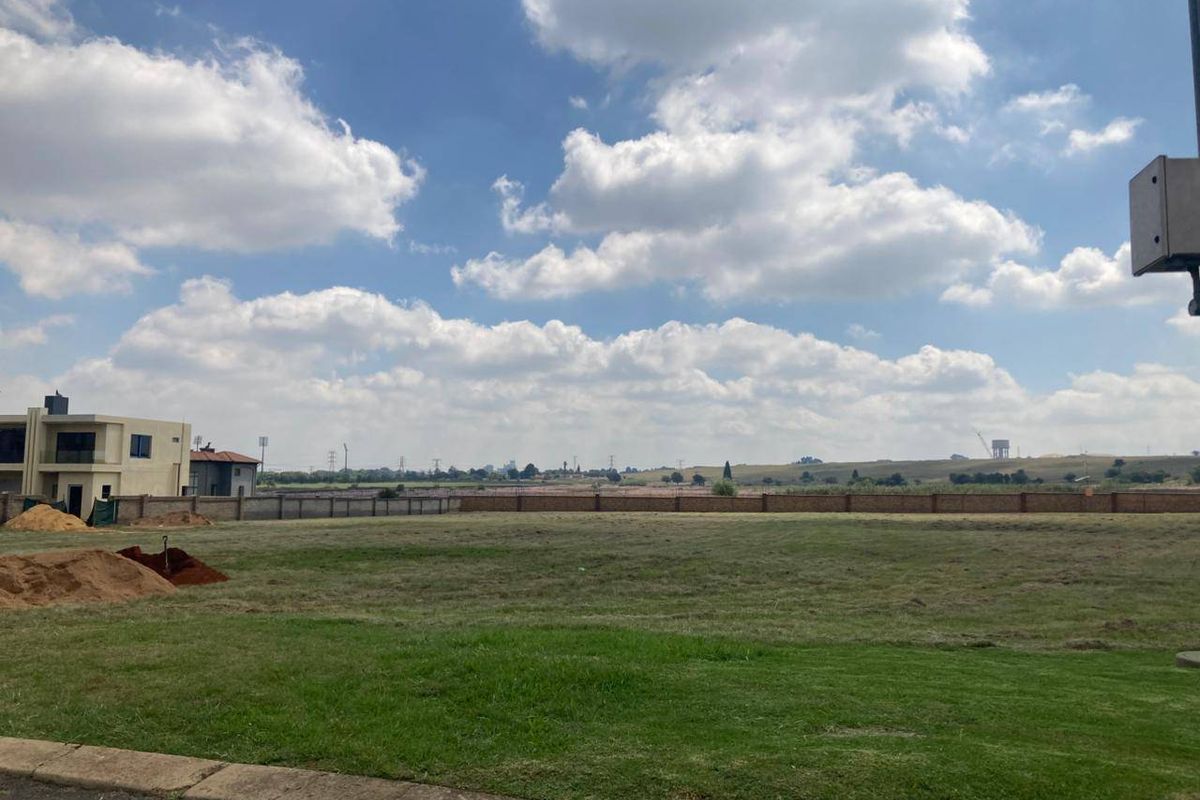 A vacant residential plot featuring a level grass lawn, perimeter brick walling, and a paved curb, offering an open view of the surrounding landscape.