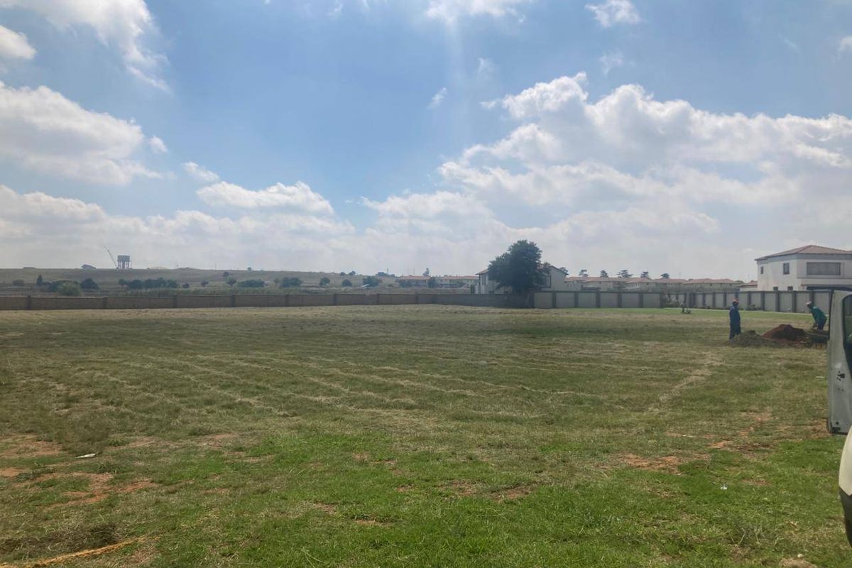 A wide, level vacant residential plot featuring a grass lawn and enclosed by a perimeter wall in a suburban setting.