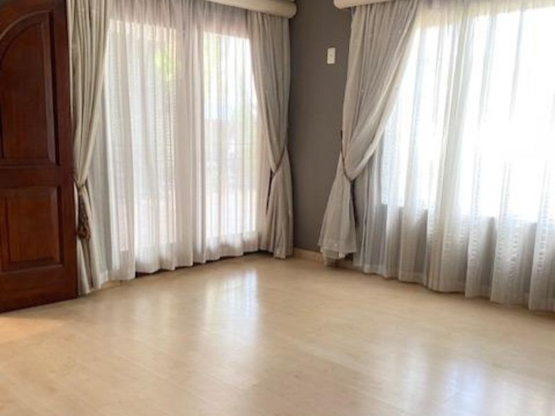 A bright, empty living room featuring light-toned laminate flooring, decorative crown molding, and large windows dressed with elegant curtains and pelmets.