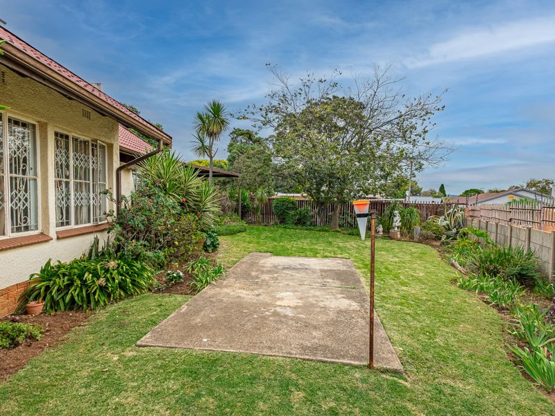 A rear garden view featuring a lawn, a concrete patio area, and a side view of the house with security bars on the windows.
