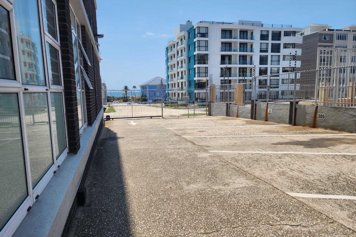 An outdoor parking area featuring concrete paving, security fencing, and numbered parking bays, situated between apartment buildings with a view towards the coast.