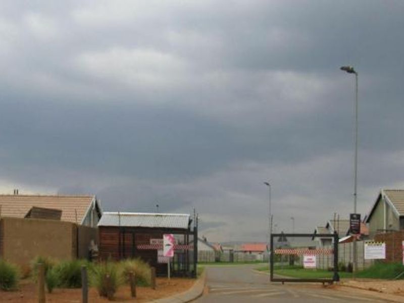 A view of the residential estate entrance featuring security gates, perimeter walls, and paved access roads under an overcast sky.