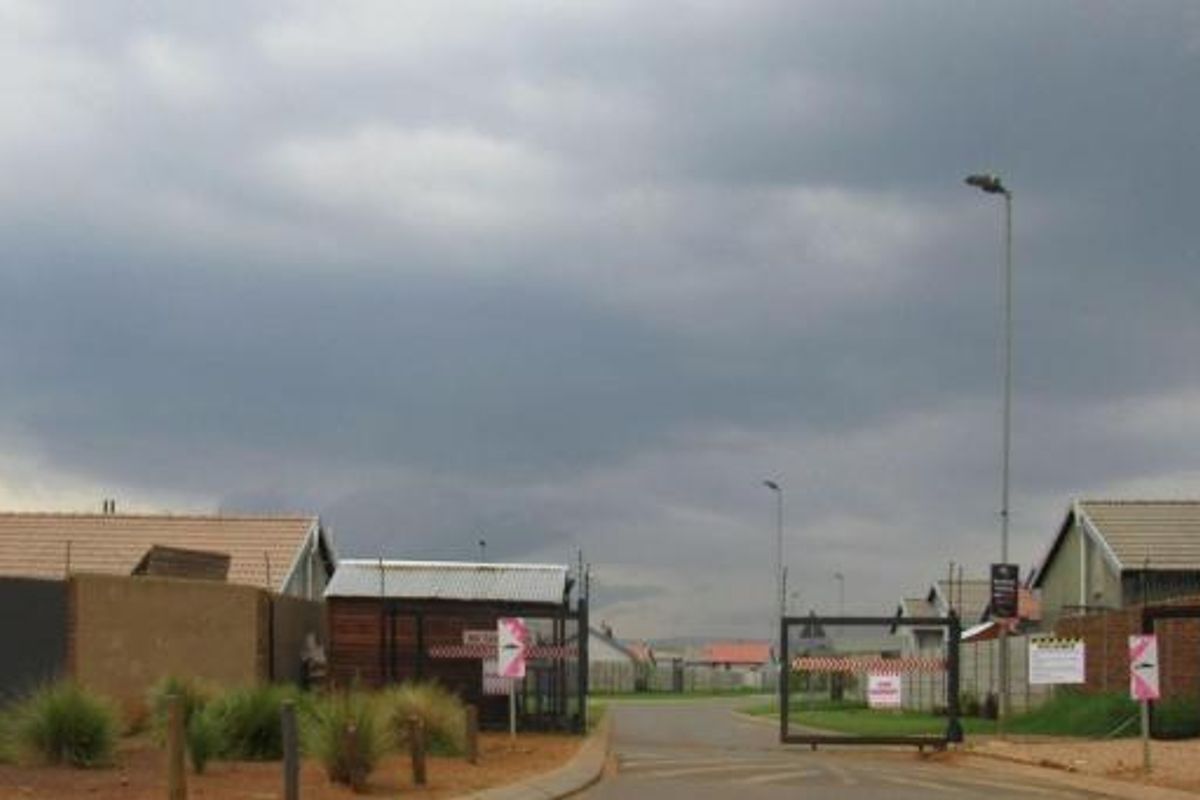 A view of the residential estate entrance featuring security gates, perimeter walls, and paved access roads under an overcast sky.