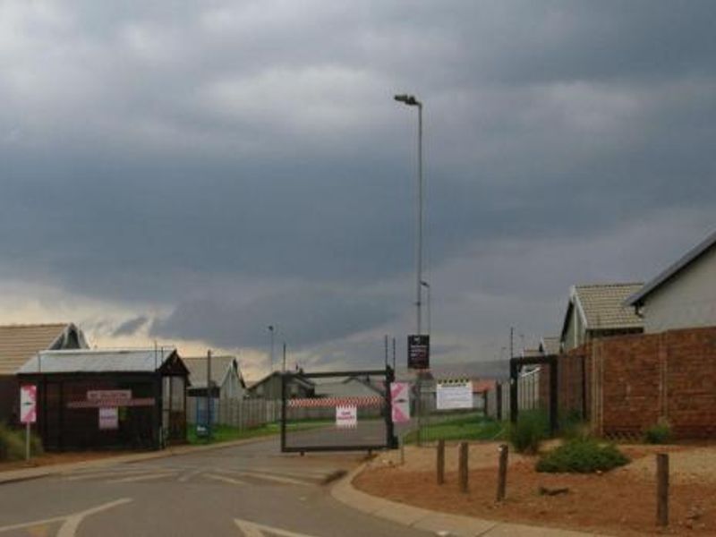 Street view of a residential estate entrance featuring a security gate, brick boundary walls with razor wire, and paved access road.