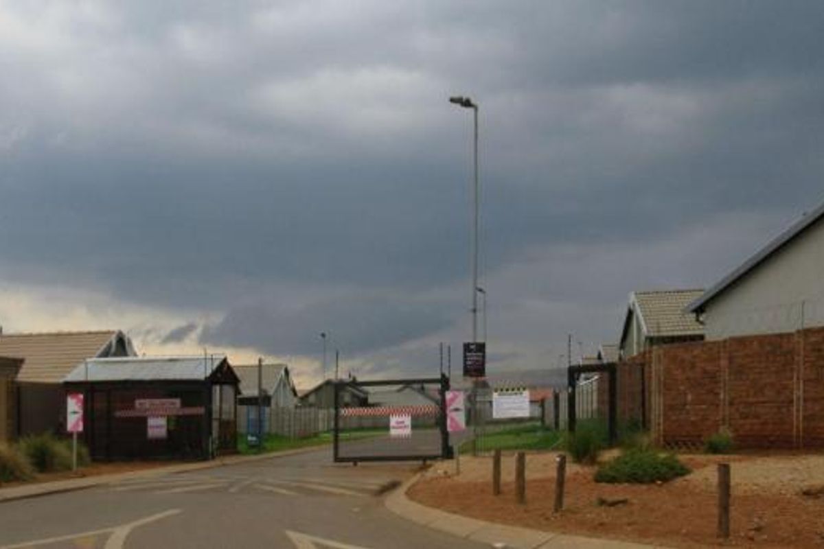 Street view of a residential estate entrance featuring a security gate, brick boundary walls with razor wire, and paved access road.