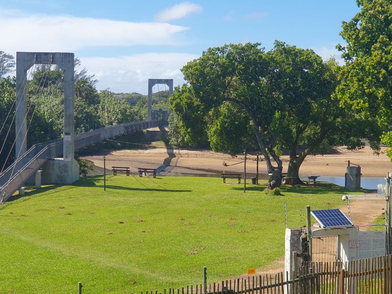 A scenic view of a suspension bridge spanning a river, set within a well-maintained green park area with security fencing and solar infrastructure.
