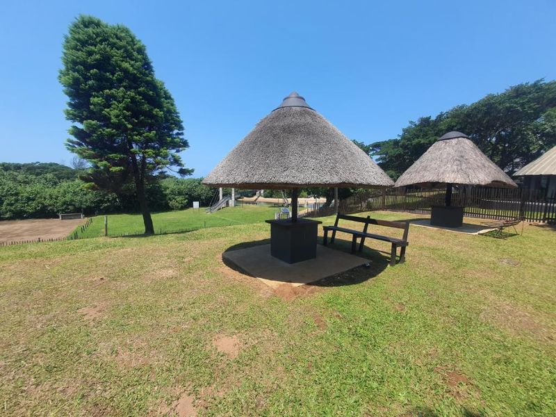 A well-maintained communal garden area featuring two traditional thatched lapas with concrete bases, set within a spacious lawned environment.