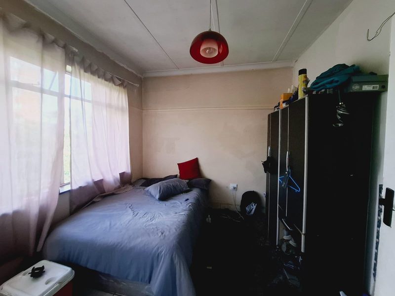 A compact bedroom featuring a large window, picture rails, and basic electrical fixtures, currently requiring cosmetic updates and wall repairs.