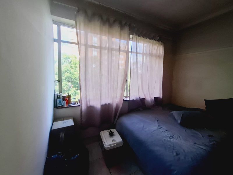 A bedroom featuring a large window providing natural light and tiled flooring, currently showing signs of wear and requiring cosmetic updates.