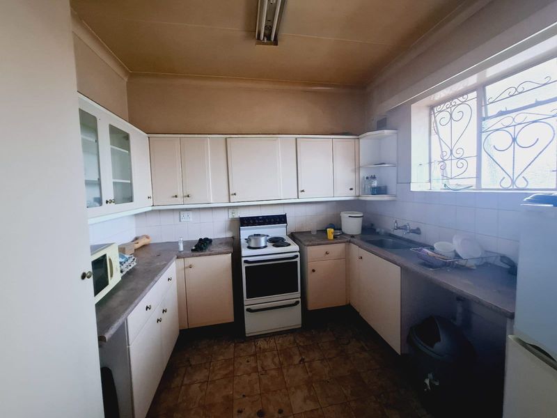 A dated kitchen featuring original cabinetry, laminate countertops, a freestanding stove, and a stainless steel sink with security bars on the window.