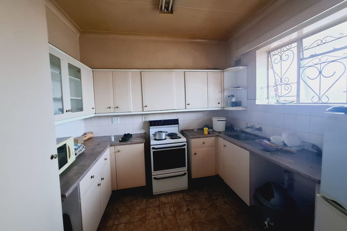 A dated kitchen featuring original cabinetry, laminate countertops, a freestanding stove, and a stainless steel sink with security bars on the window.