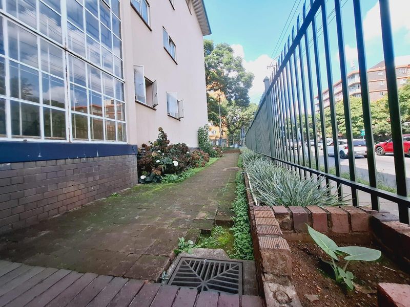 A side exterior walkway featuring brick paving, a metal security fence, and a built-in brick planter box alongside the apartment building facade.