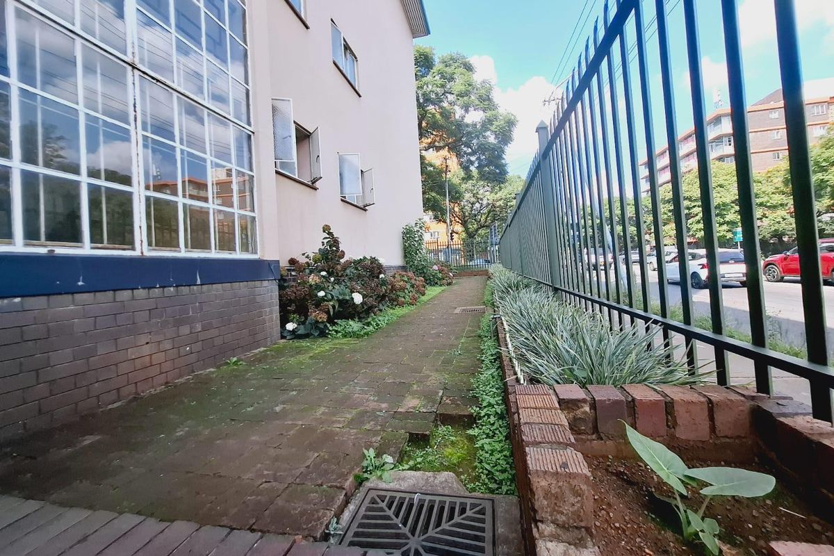 A side exterior walkway featuring brick paving, a metal security fence, and a built-in brick planter box alongside the apartment building facade.