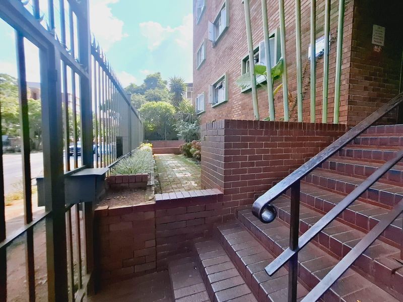 Exterior view of the apartment building entrance featuring a brick facade, paved walkway, and secure metal fencing.