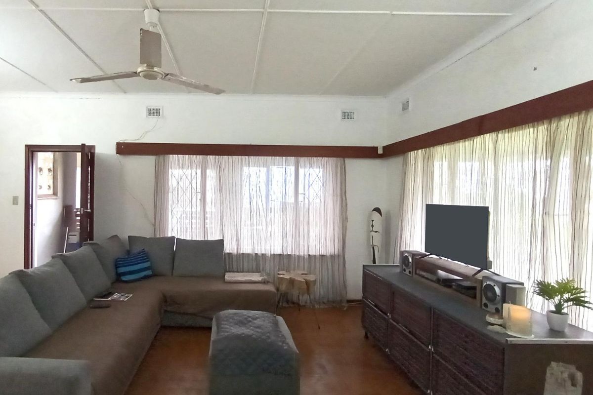 A spacious living room featuring a ceiling fan, timber curtain pelmets, and large windows with security gates, offering a functional and well-lit living space.