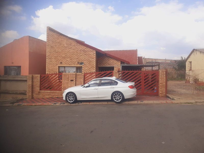Front elevation of a face brick house featuring a pitched tiled roof, secure metal gate, and paved driveway area.
