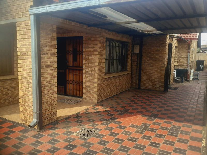 Front entrance area featuring a face brick facade, paved driveway, and a covered carport structure with security gate access.