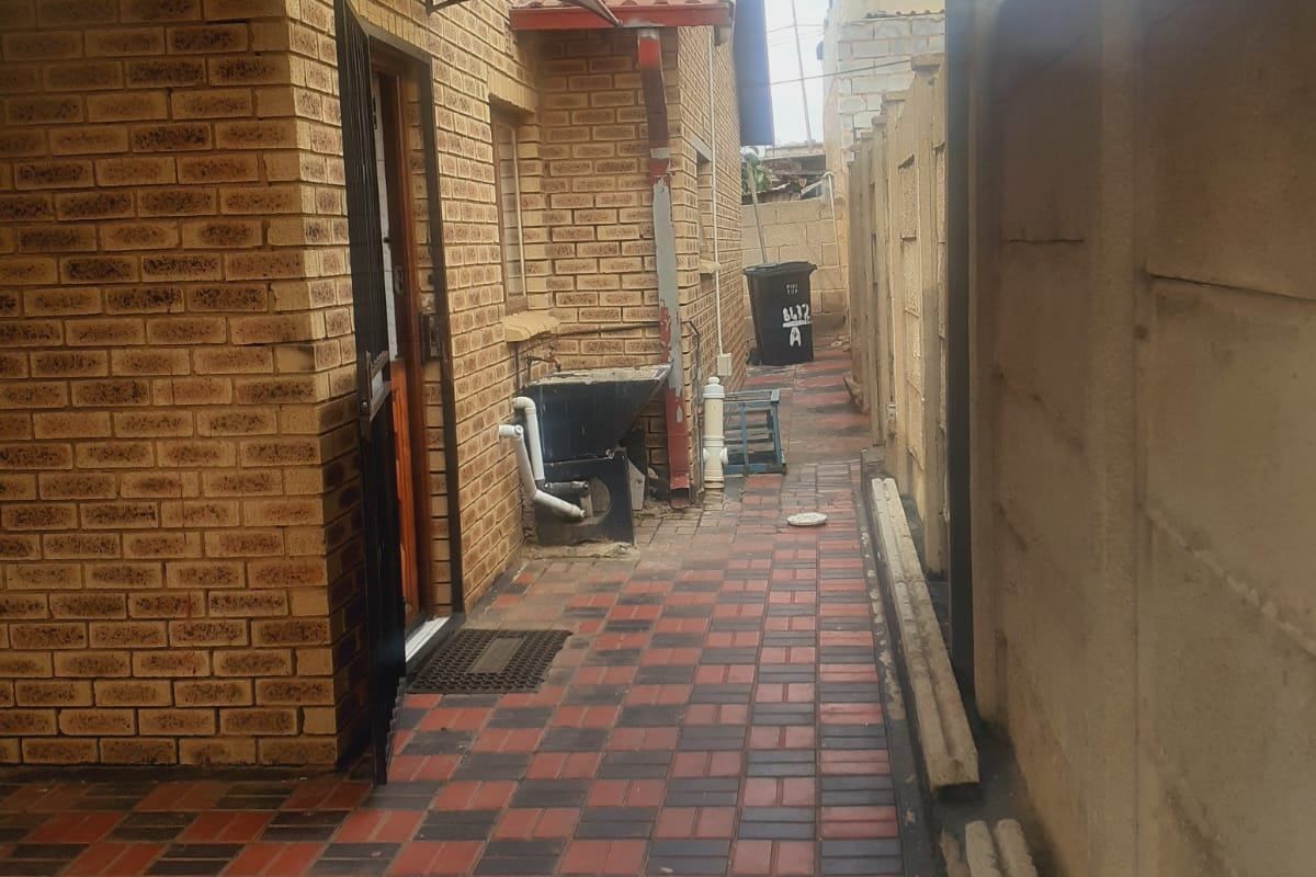 A side exterior walkway featuring brick wall construction, paved flooring, and a covered patio area leading to the rear of the property.