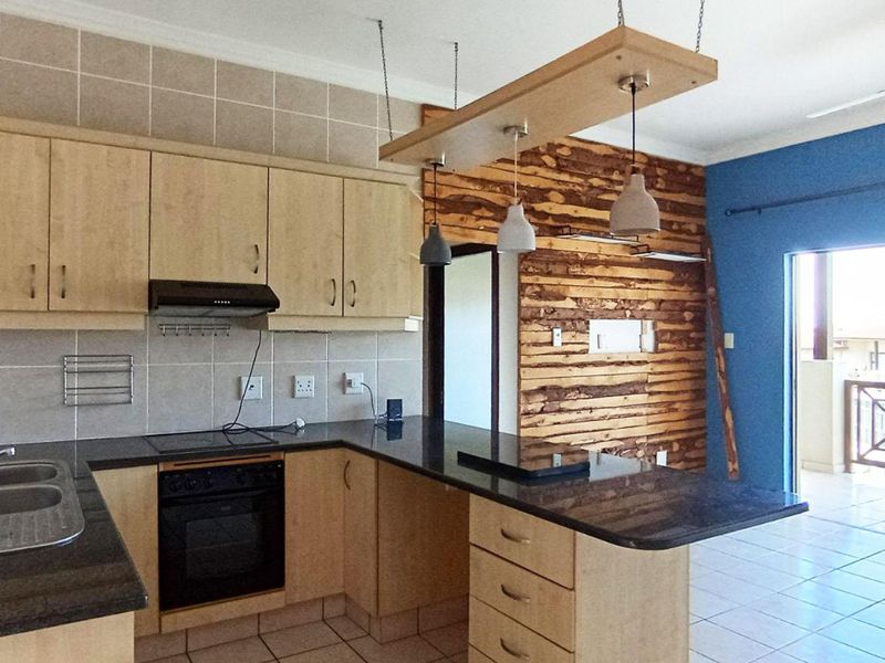 Open-plan kitchen featuring light wood cabinetry, granite countertops, and a unique timber feature wall, flowing into a living area with access to a balcony.