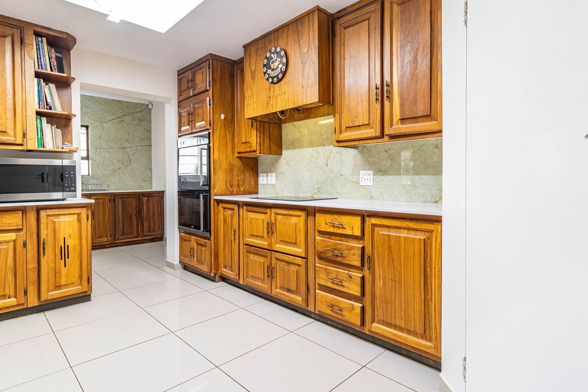 A functional kitchen featuring extensive wooden cabinetry, built-in oven, integrated hob, and light-colored floor tiles, with an adjacent scullery area.
