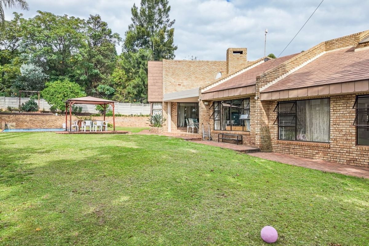 Spacious rear garden featuring a brick-faced house, covered patio area, and established lawn with mature trees and perimeter walling.