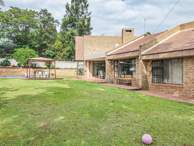 Spacious rear garden featuring a brick-faced house, covered patio area, and established lawn with mature trees and perimeter walling.