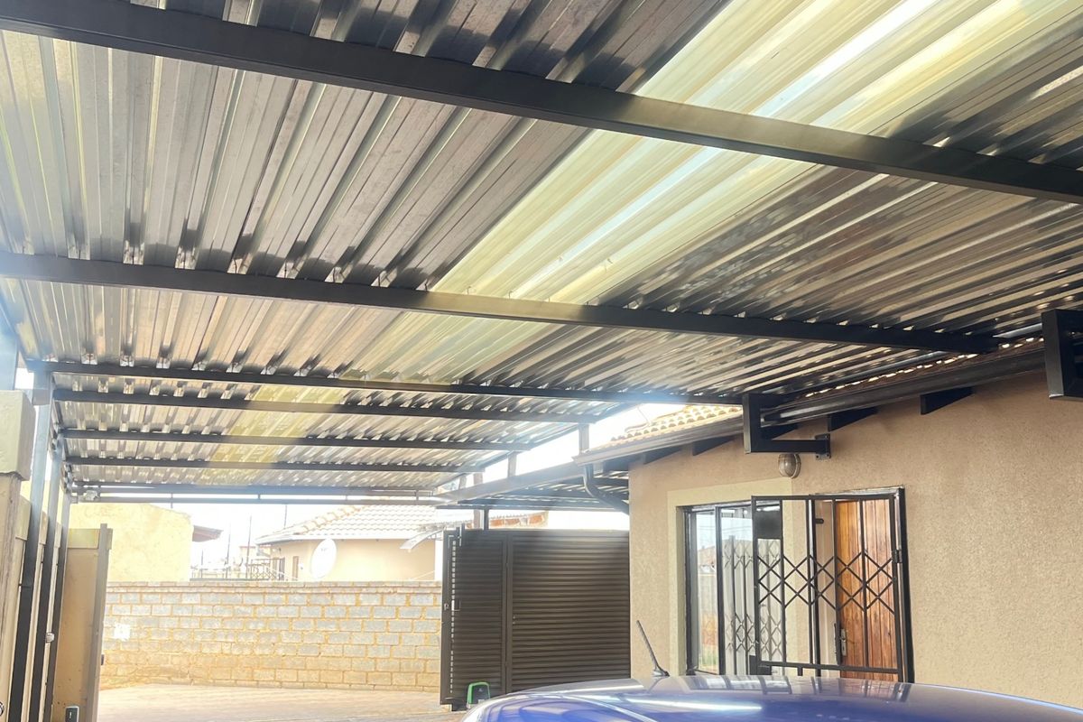 A covered carport area featuring a corrugated metal roof with steel support beams, paved driveway, and secure access gate.