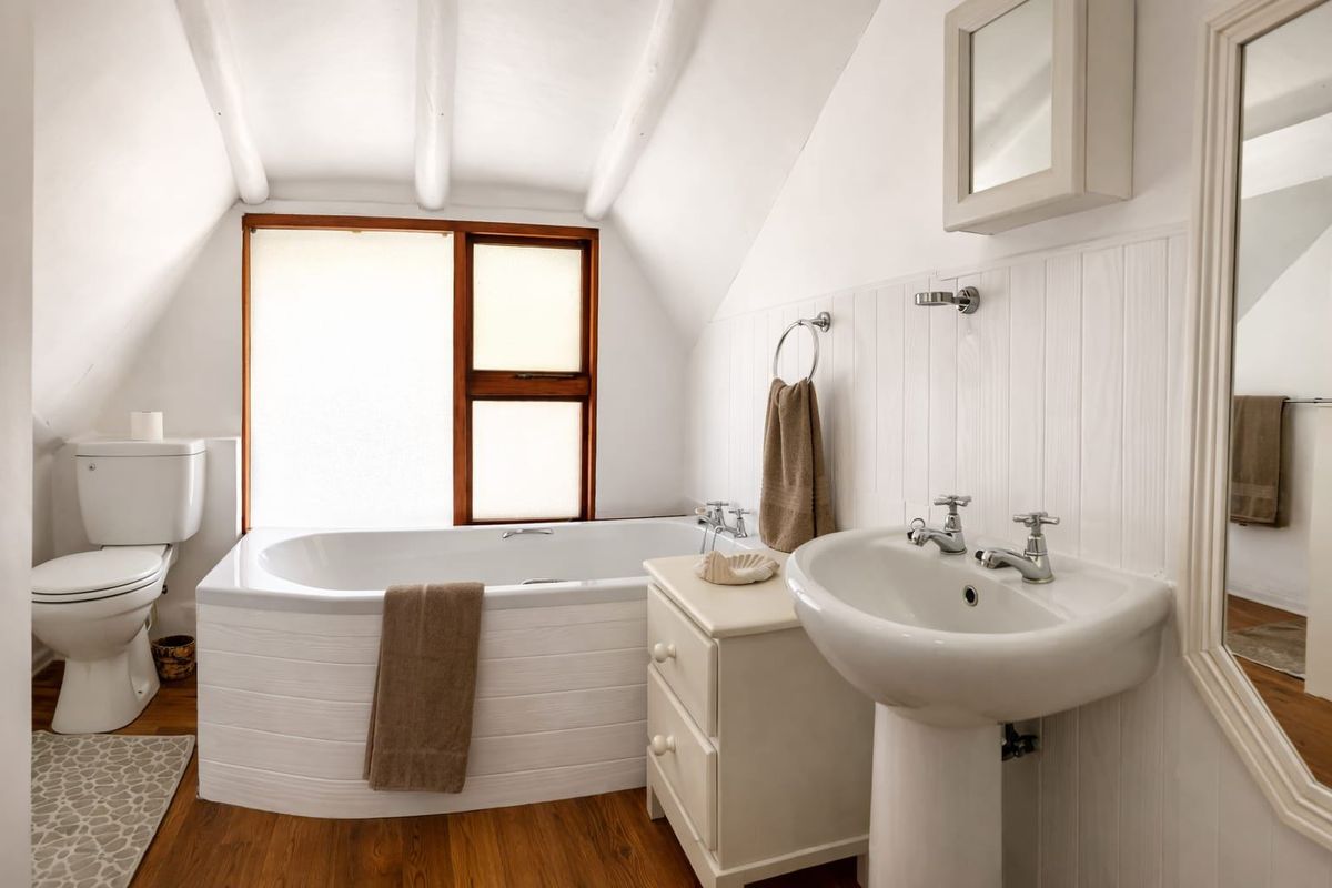 A bright bathroom featuring a pedestal basin, bathtub, toilet, and charming exposed ceiling beams with timber window frames and wall paneling.