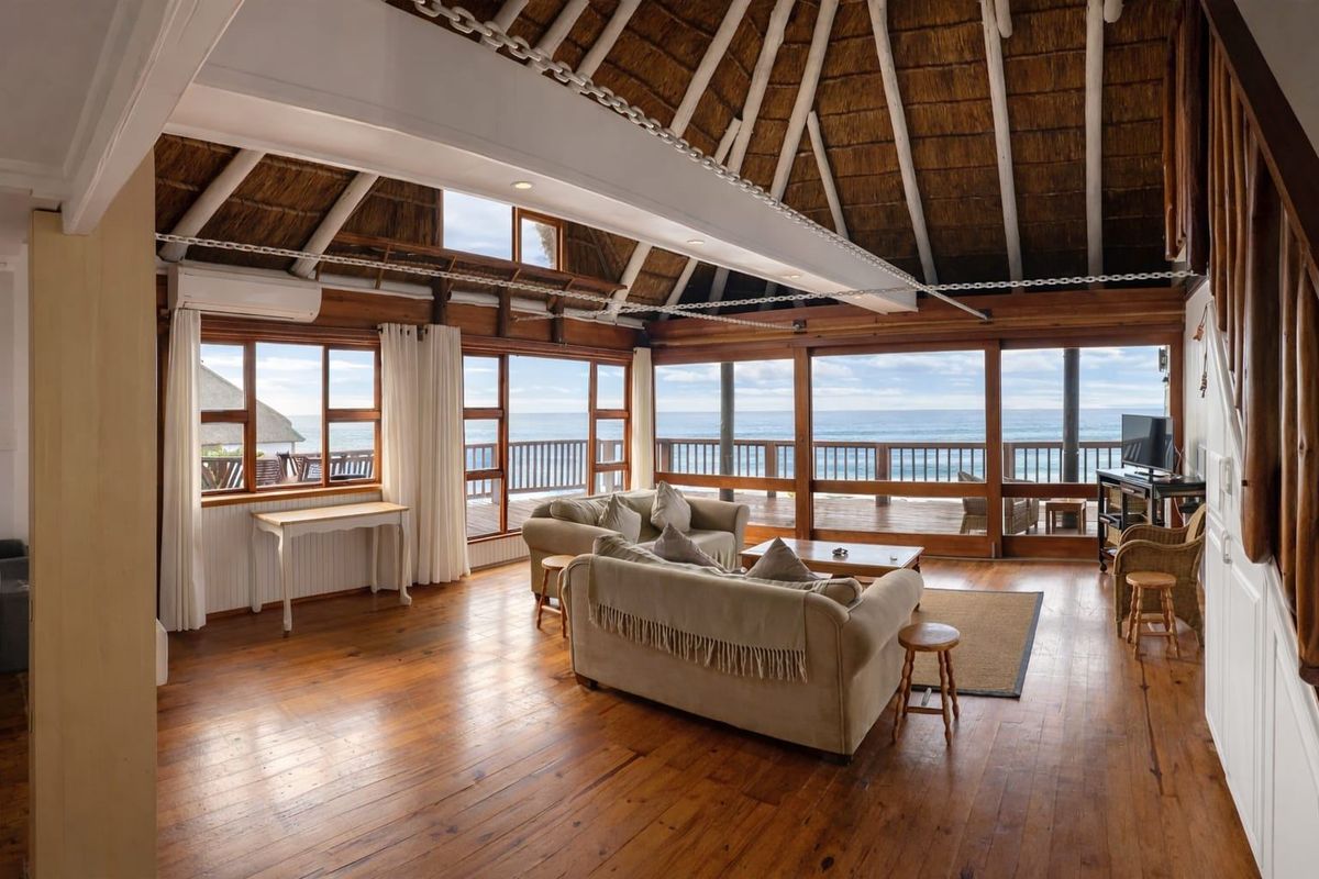 Spacious living area featuring a high thatched ceiling with exposed beams, warm timber flooring, and expansive windows offering panoramic ocean views and deck access.