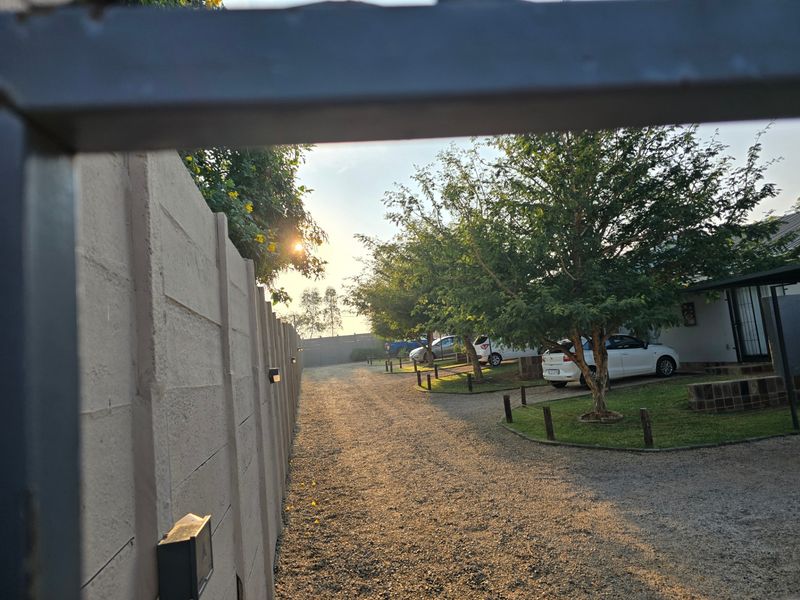 A view of the property entrance featuring a gravel driveway, precast boundary wall, and landscaped garden area with mature trees.