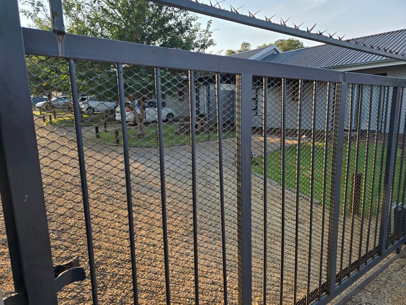 Front view of the property featuring a secure metal gate with spiked topping, gravel driveway, and a metal-roofed building structure.