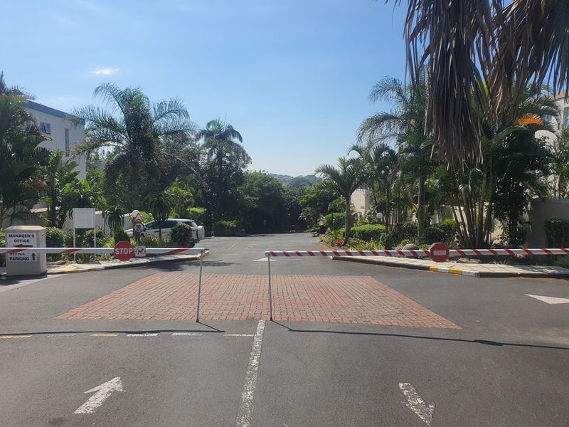 View of the secure entrance to the apartment complex featuring a boom gate, paved driveway, and established landscaped gardens.