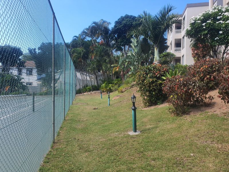 A well-maintained tennis court area featuring a chain-link perimeter fence, landscaped garden borders, and integrated pathway lighting.