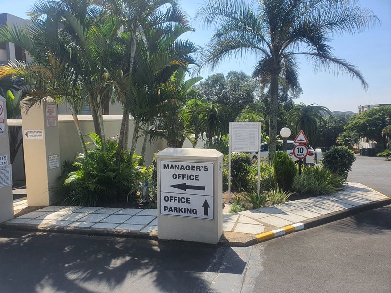 Entrance area of the apartment complex featuring paved driveway, landscaped gardens, and clear directional signage for the manager's office and parking.