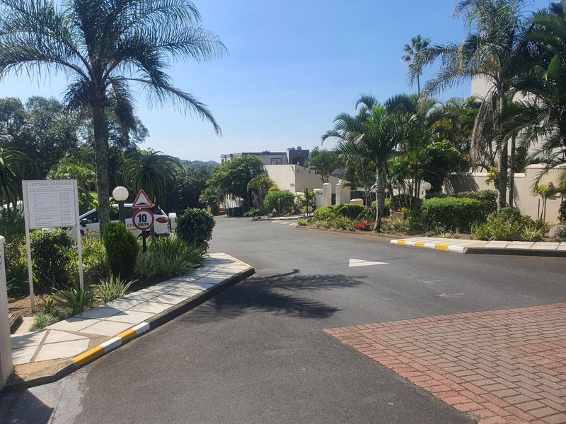 A view of the complex entrance and access road, featuring well-maintained landscaping, palm trees, and paved driveway infrastructure.