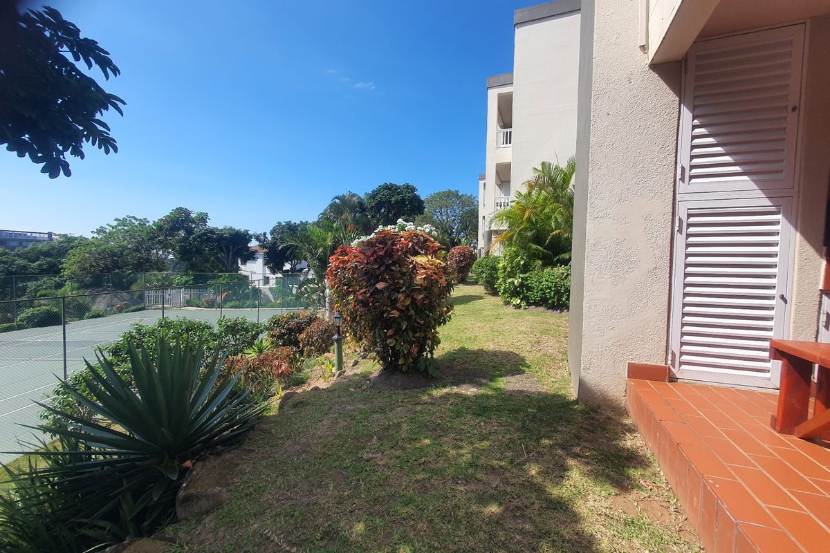 A view of the rear exterior featuring a tiled patio area, established garden landscaping, and adjacent tennis court facilities.