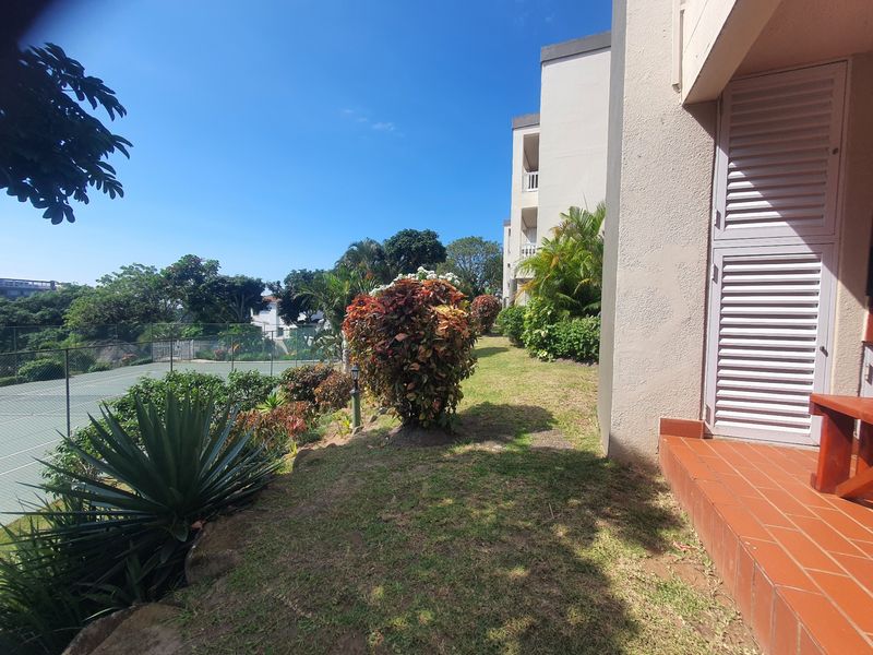 A view of the rear exterior featuring a tiled patio area, established garden landscaping, and adjacent tennis court facilities.