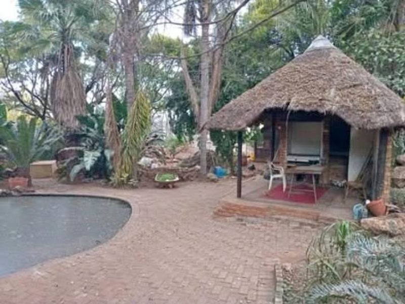 Outdoor pool area featuring a traditional thatched lapa structure and brick paved surround, set within a mature, heavily landscaped garden environment.