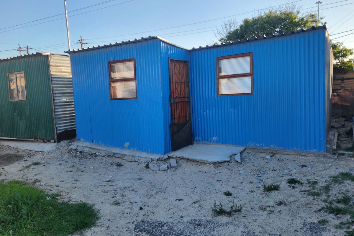 A small, blue corrugated metal structure featuring a security gate and concrete foundation, situated on an unpaved residential plot.