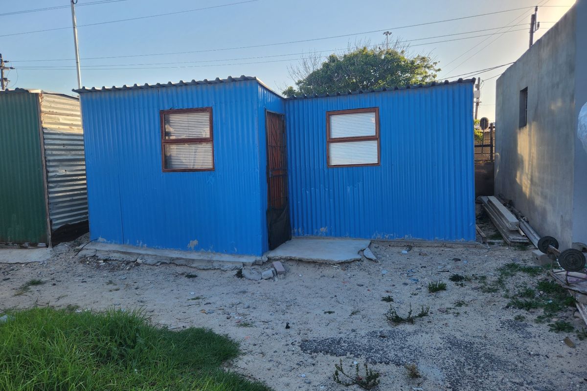 A small, blue corrugated metal structure featuring a concrete slab foundation and security gate, situated on an unpaved residential plot.