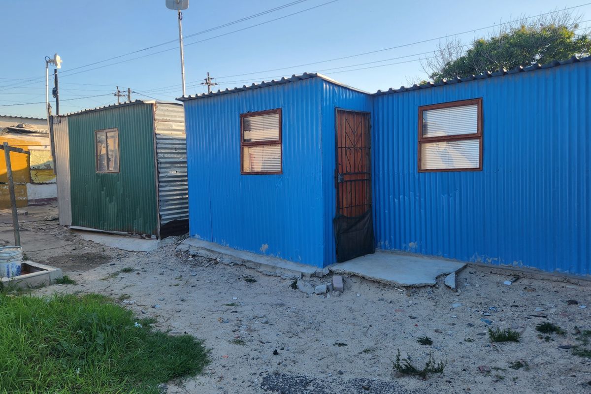 Exterior view of two small corrugated metal structures on a sandy residential plot with basic security gates and concrete foundations.