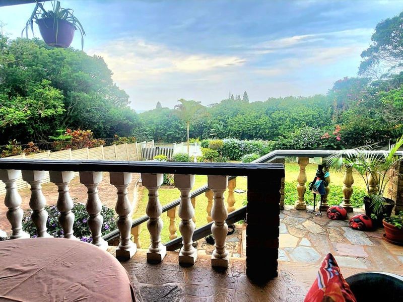 A covered patio featuring stone flooring and a classic concrete balustrade, overlooking a lush, green garden and surrounding trees.