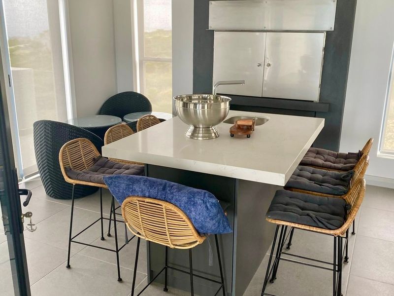 Modern covered patio area featuring a built-in braai, a central stone-topped island with an integrated sink, and neutral tiled flooring.