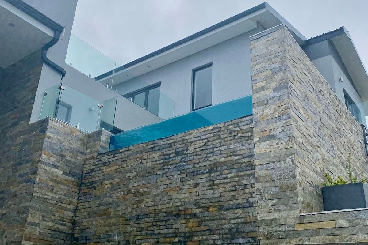 A modern architectural exterior featuring a striking elevated glass-walled swimming pool and premium stone cladding facade.