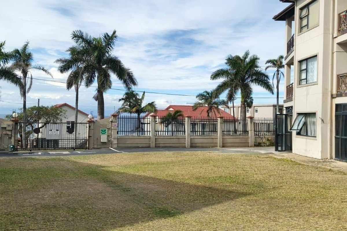 A view of the apartment complex exterior featuring a lawn area, perimeter security fencing, and multi-level balconies.