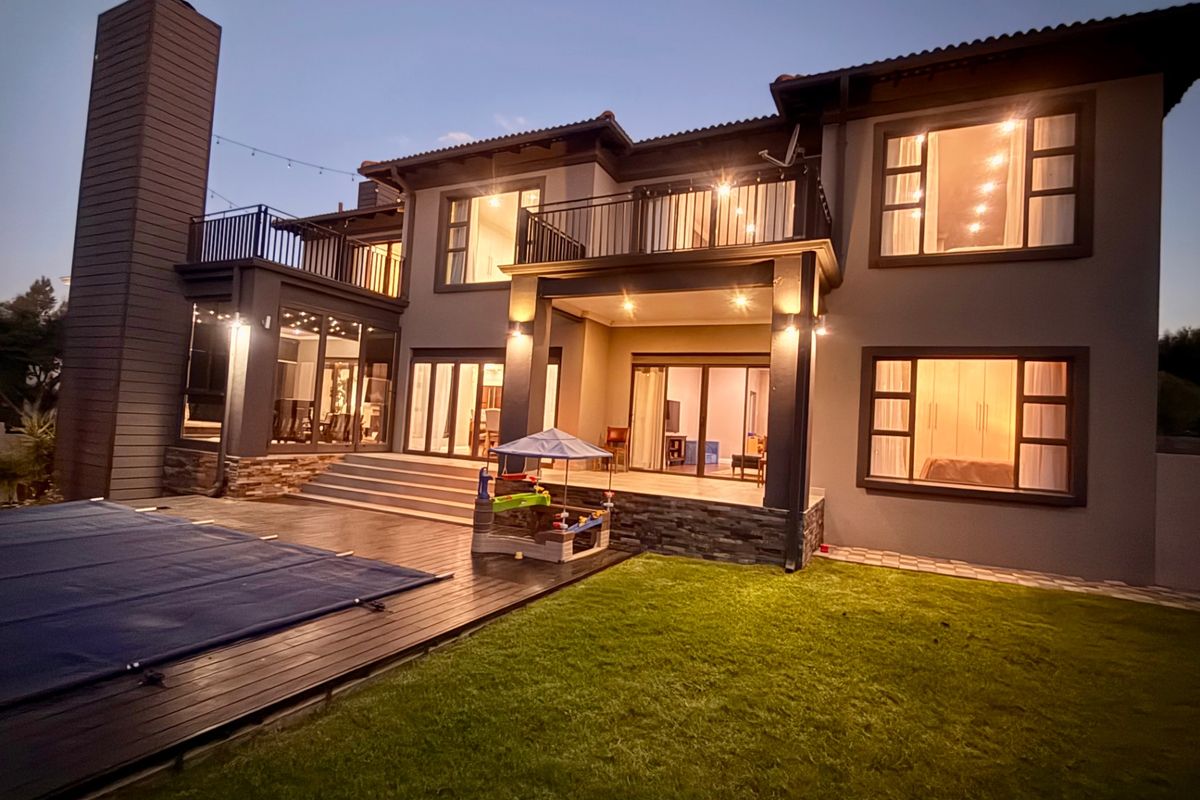 Rear elevation of a two-story house featuring a swimming pool with cover, wooden decking, a balcony, and stone-clad accents under evening lighting.