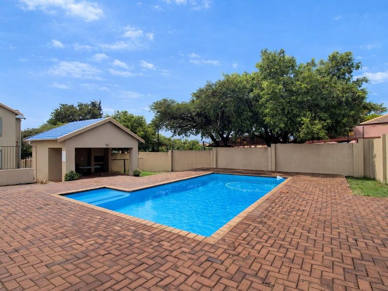 A well-maintained communal swimming pool area featuring paved decking and a covered entertainment structure with a built-in braai.