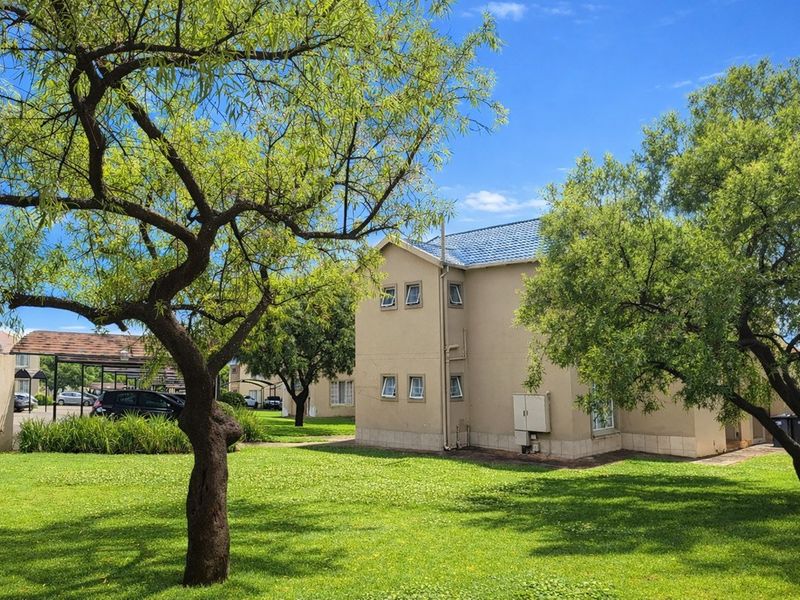 A view of the apartment complex exterior featuring well-maintained lawns, mature trees, and a tiled roof structure with adjacent carport parking.