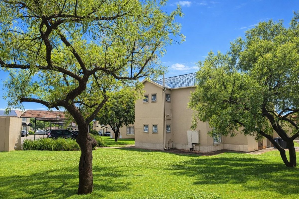 A view of the apartment complex exterior featuring well-maintained lawns, mature trees, and a tiled roof structure with adjacent carport parking.